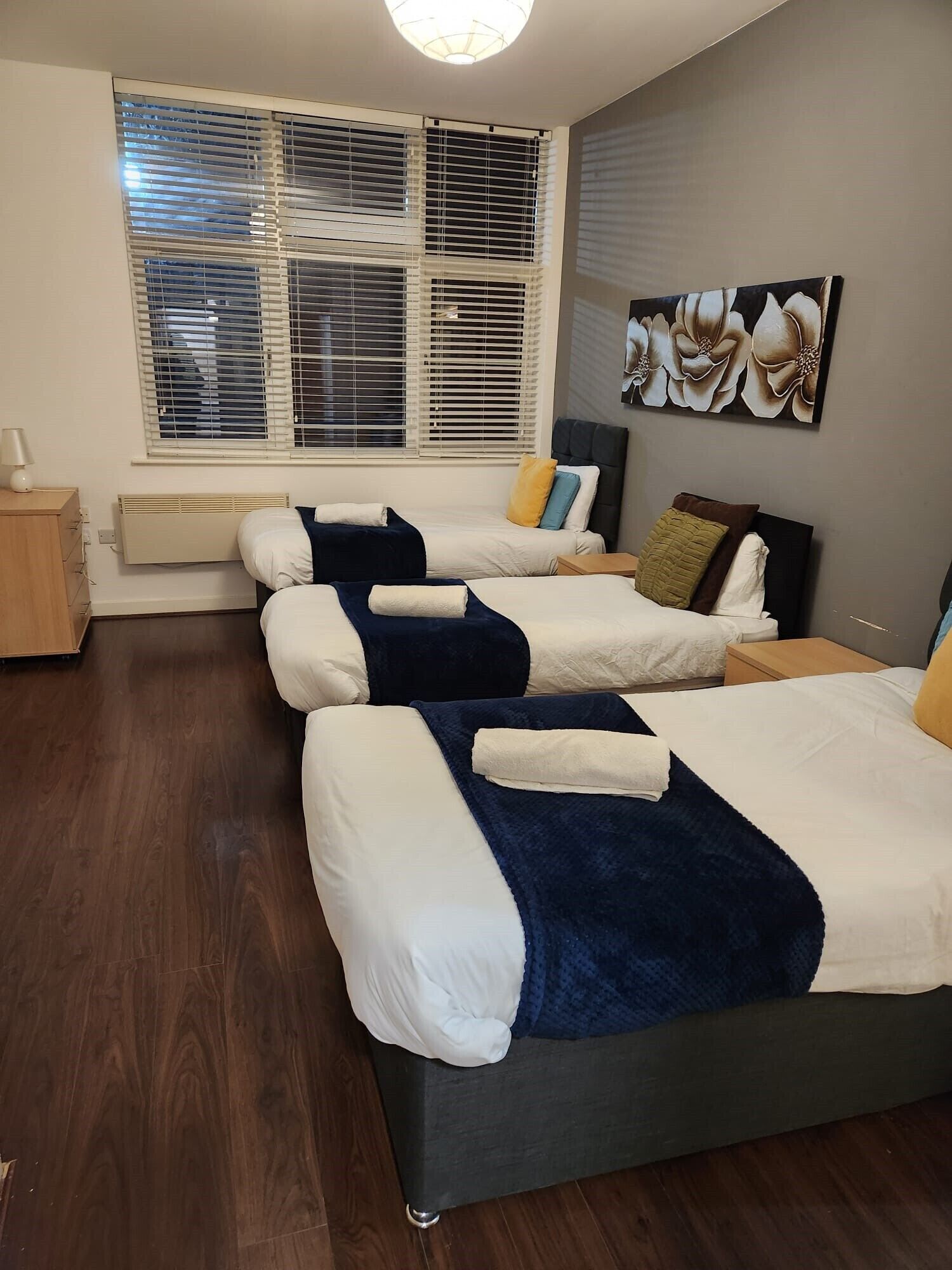 A hotel room with two beds, a large window with blinds, a radiator, a bedside table, a lamp, and a wall-mounted artwork.
