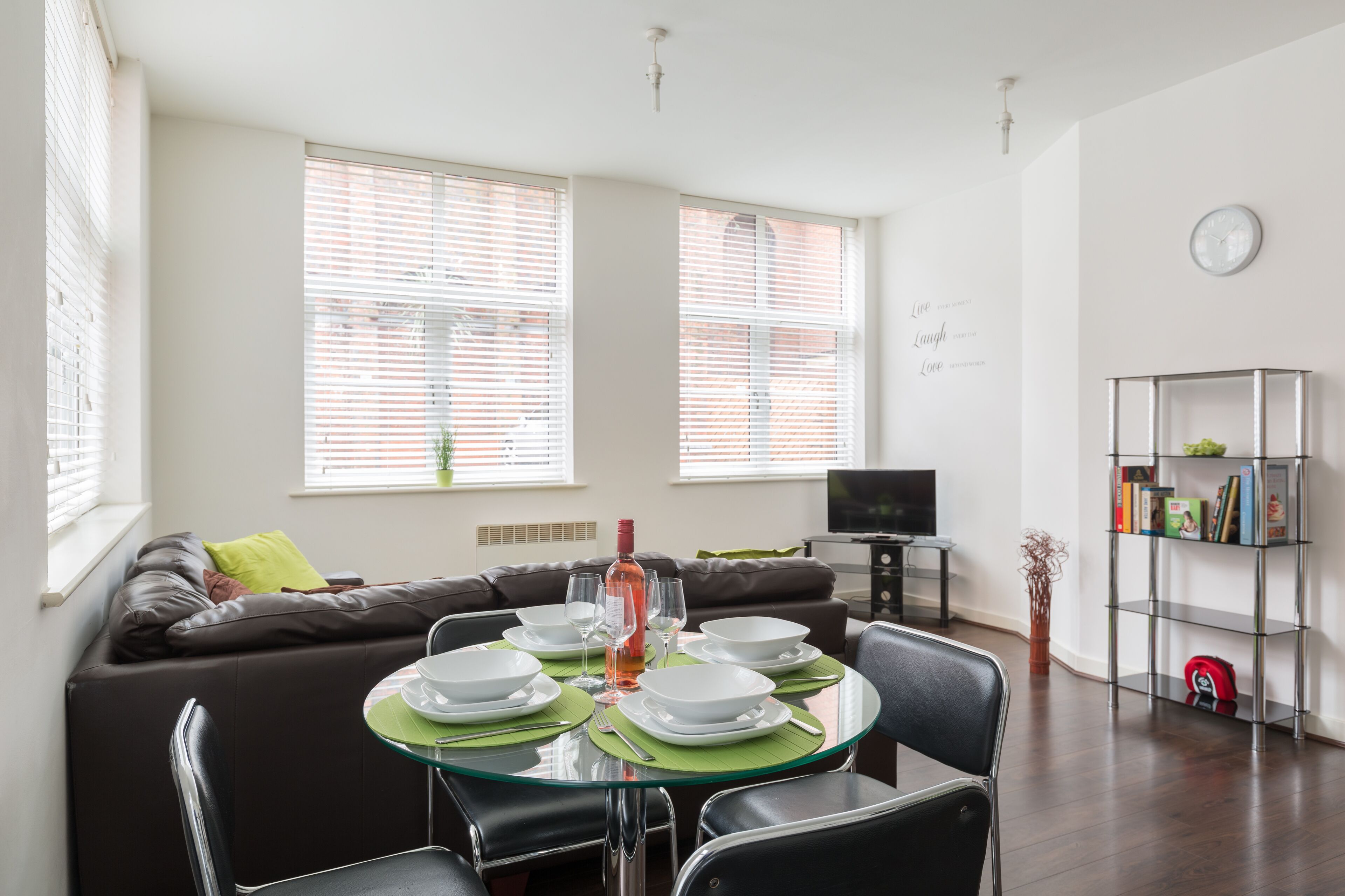 A dining area with a round glass table set for four, surrounded by black chairs. There is a brown leather sofa with a green pillow and a brown throw blanket in the background. A television is mounted on the wall to the right.