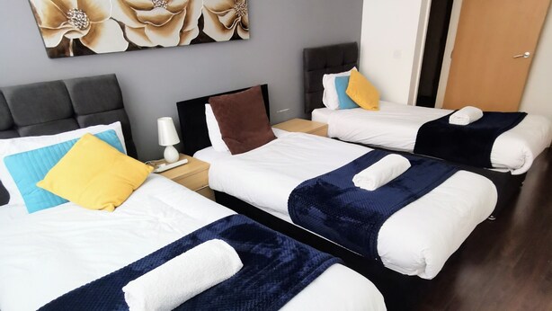 A hotel room with two beds, each with a blue and white duvet, a brown pillow, and a white pillow. There is a nightstand with a lamp and a small table. A large floral painting is on the wall above the beds. - 2bed Apt -king Bed -free Parking - Family Friendly (Birmingham)