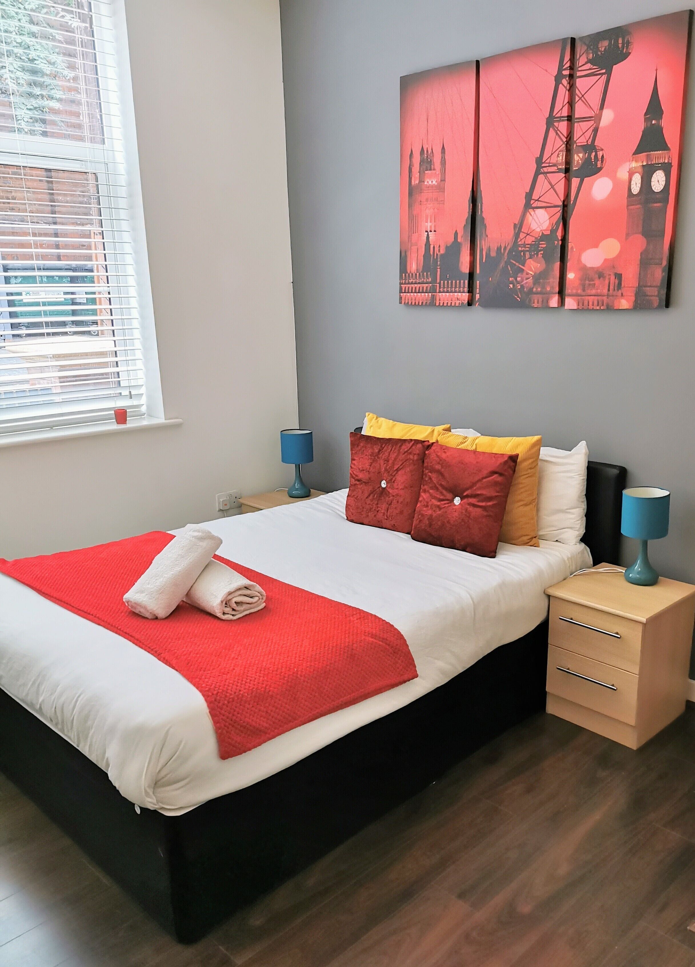 A neatly made bed with a red blanket and pillows, a bedside table with a lamp, and a wall with a cityscape painting.