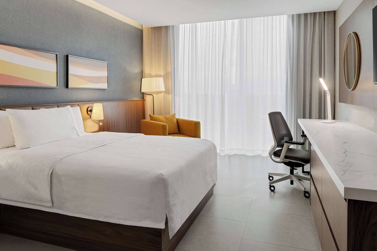 A hotel room with a bed, a desk, a chair, and a large window with curtains.