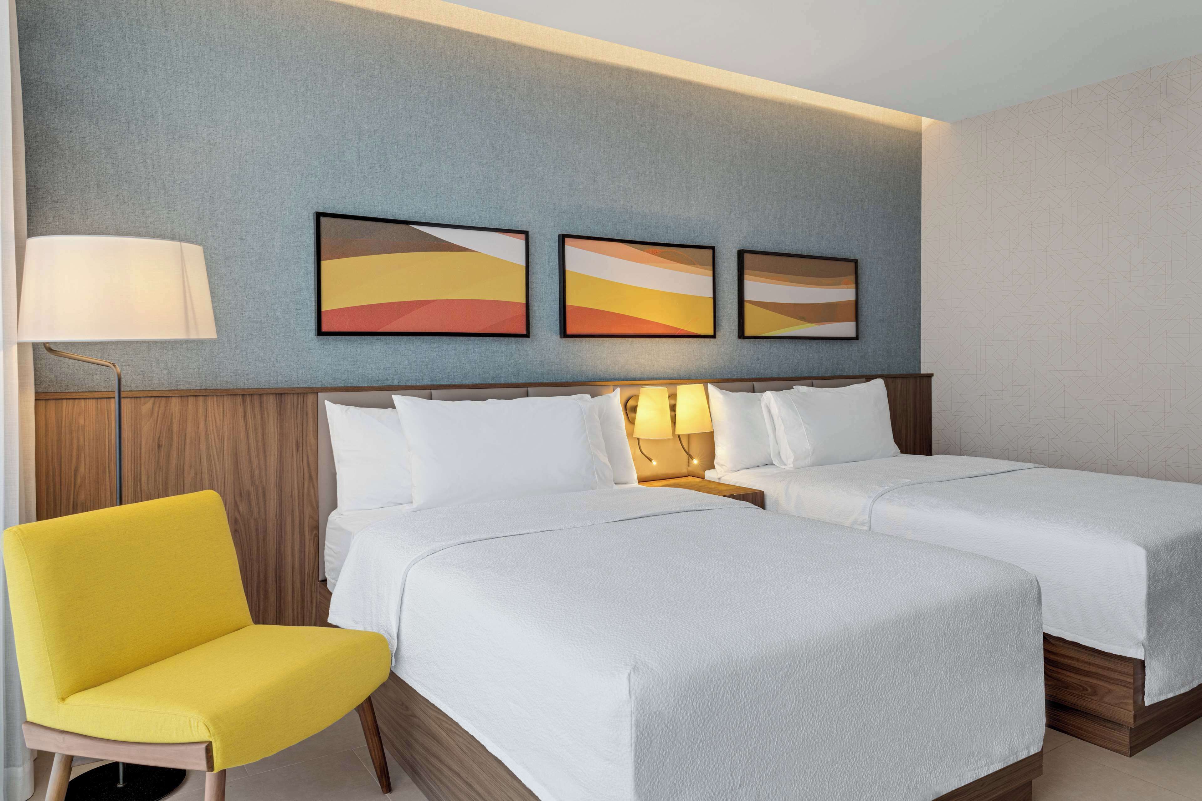 A hotel room with two beds, a yellow chair, and three abstract paintings on the wall.