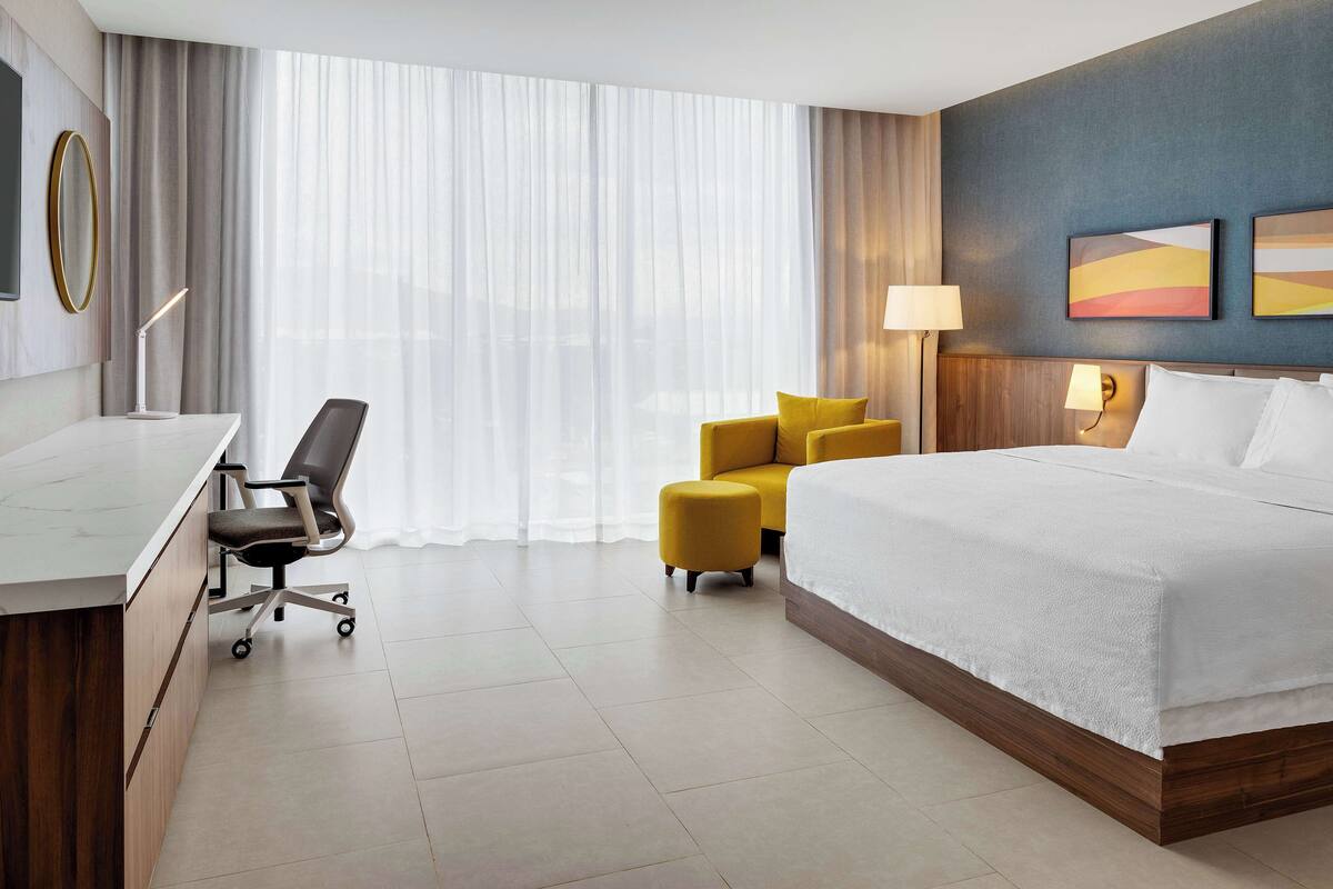 A hotel room with a bed, a desk, a chair, a small ottoman, and a large window with curtains.