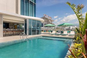 Un área de piscina con sillones de descanso y sombrillas, un bar y un cartel que dice 'City Mall'.