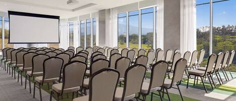 A conference room with rows of chairs, a large screen, and large windows offering a view of the outdoors.