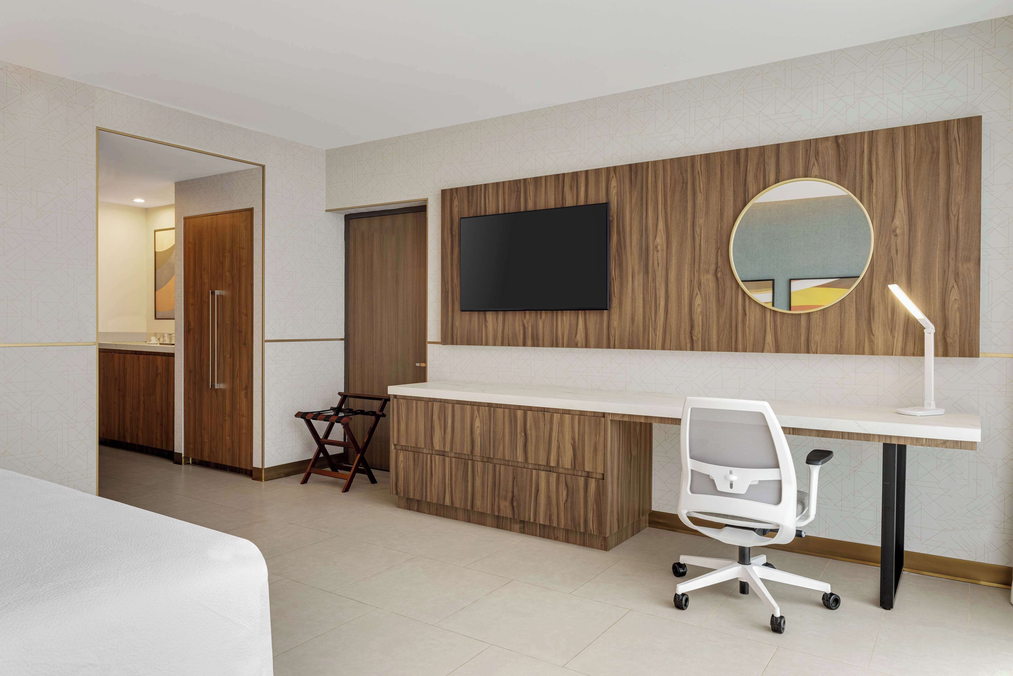 Una habitación de hotel moderna con un escritorio de madera, un televisor de pantalla plana, un espejo redondo y una silla de oficina blanca.