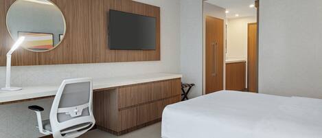 A modern hotel room with a wooden desk, a white office chair, a large bed, a round mirror, and a flat-screen TV mounted on the wall.