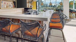 A modern bar area with wicker chairs, a bar counter, and shelves with bottles. - Hilton Garden Inn San Jose Airport City Mall (Alajuela)