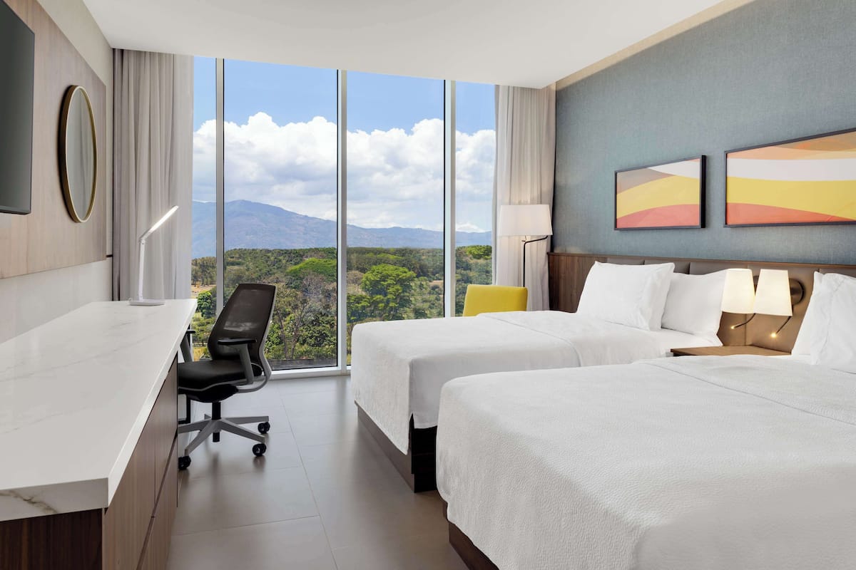 A hotel room with two beds, a desk, and a chair. There are large windows offering a view of mountains and greenery.