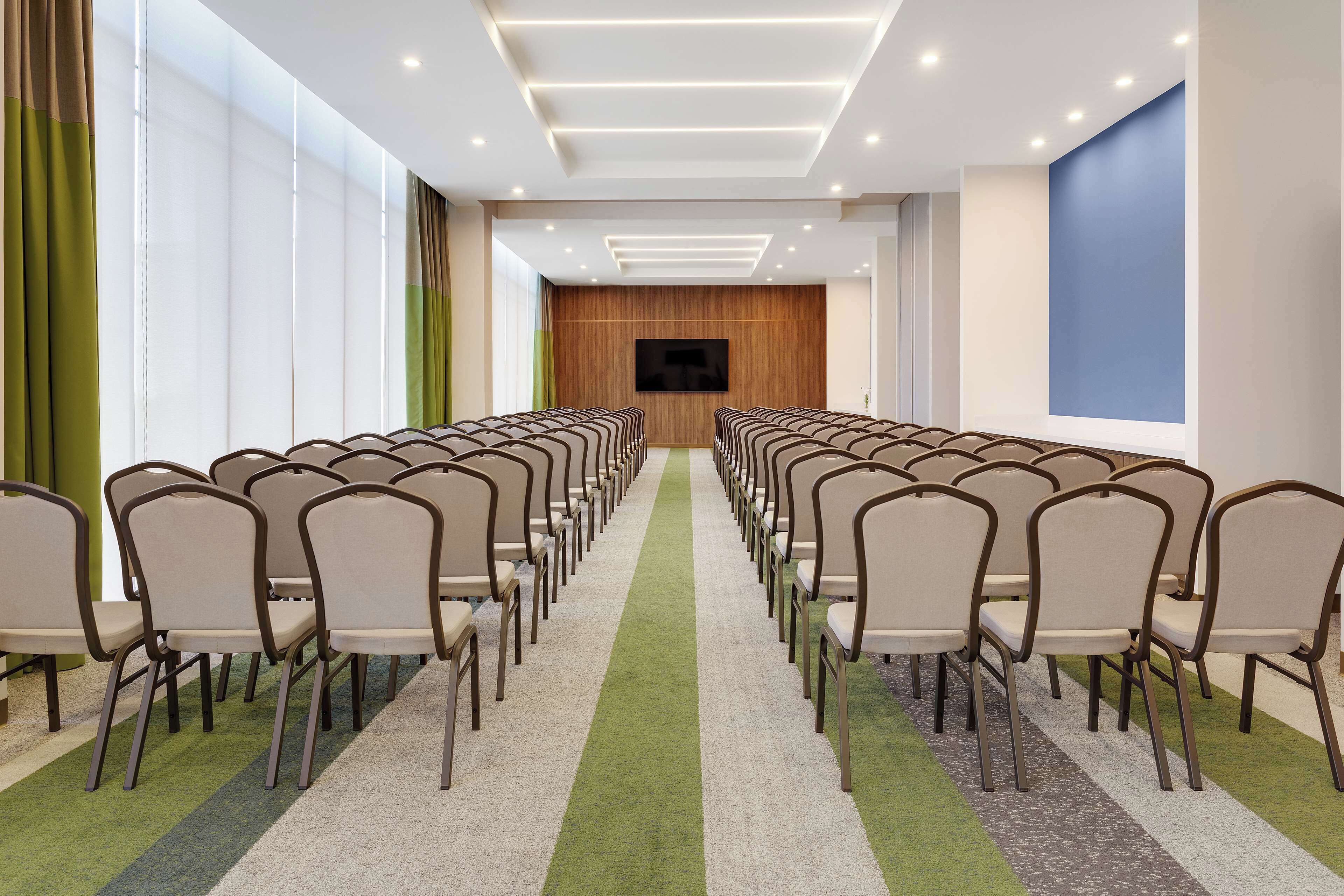 Un salón de conferencias con filas de sillas, una pantalla grande y piso alfombrado.