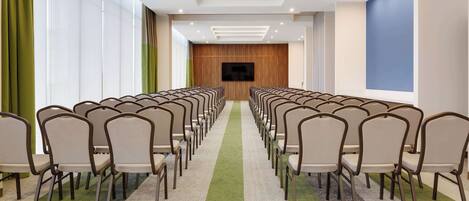 Un salón de conferencias con filas de sillas, una pantalla grande y piso alfombrado.