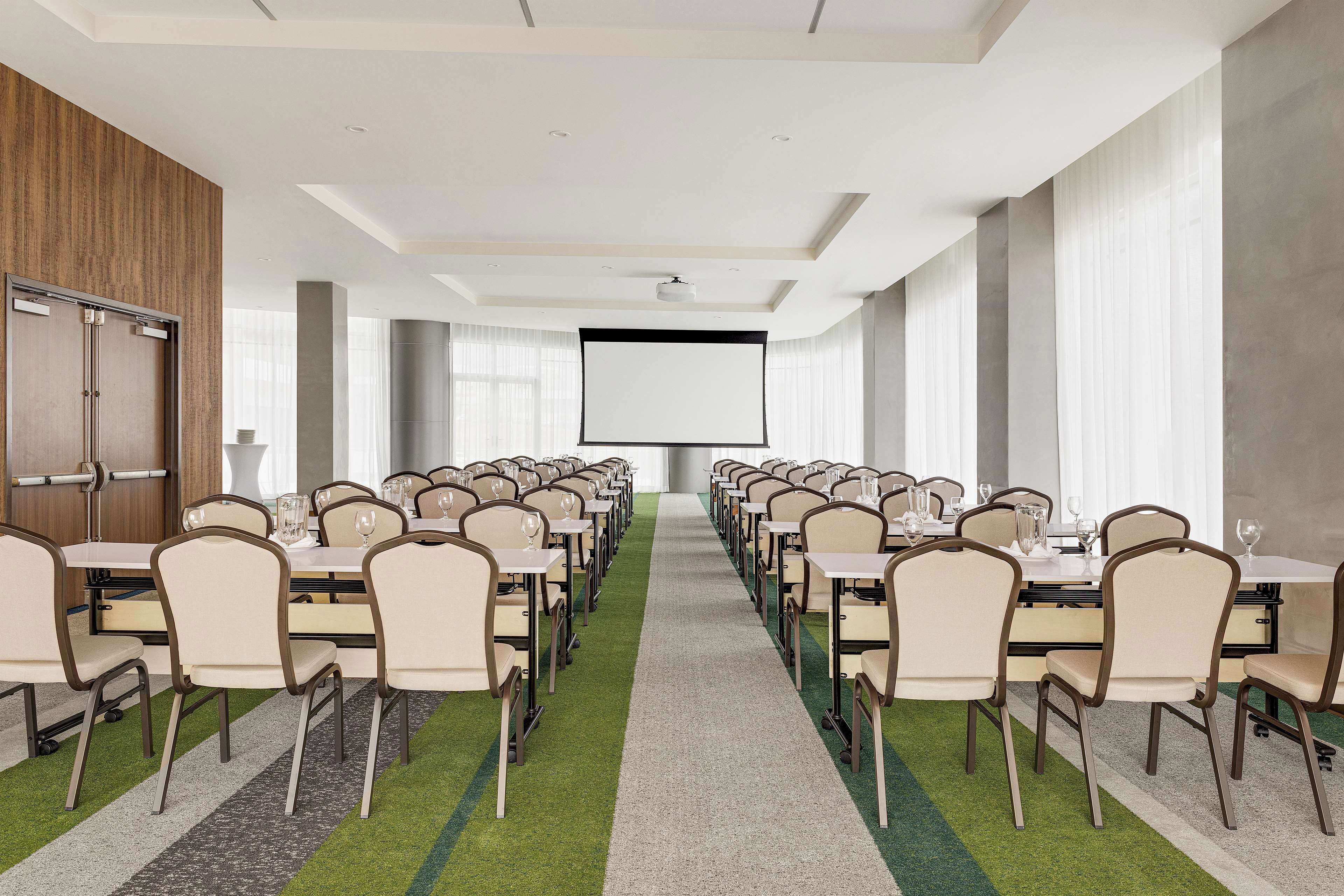 A conference room with rows of tables and chairs, a projector screen, and a carpeted floor.