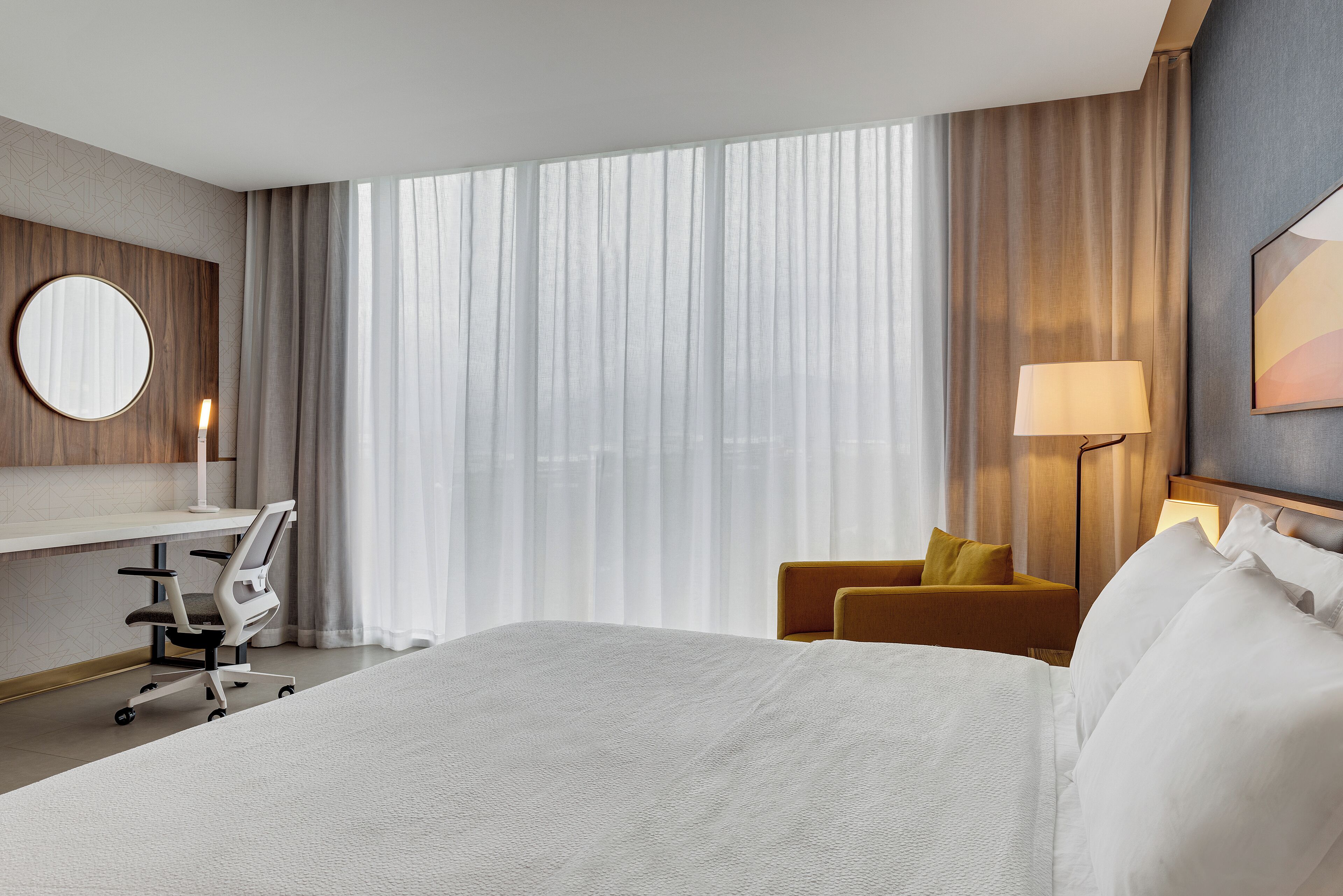 A hotel room with a large bed, a desk with a chair, a lamp, and a view of the outdoors through sheer curtains.