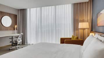 A hotel room with a large bed, a desk with a chair, a lamp, and a view of the outdoors through sheer curtains.