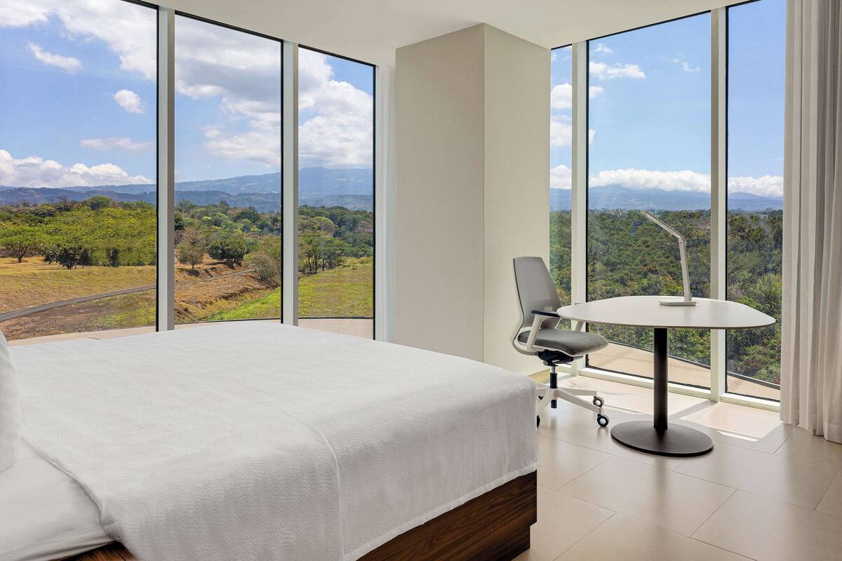 A bedroom with a large bed, a round table, and a view of a green landscape through large windows.