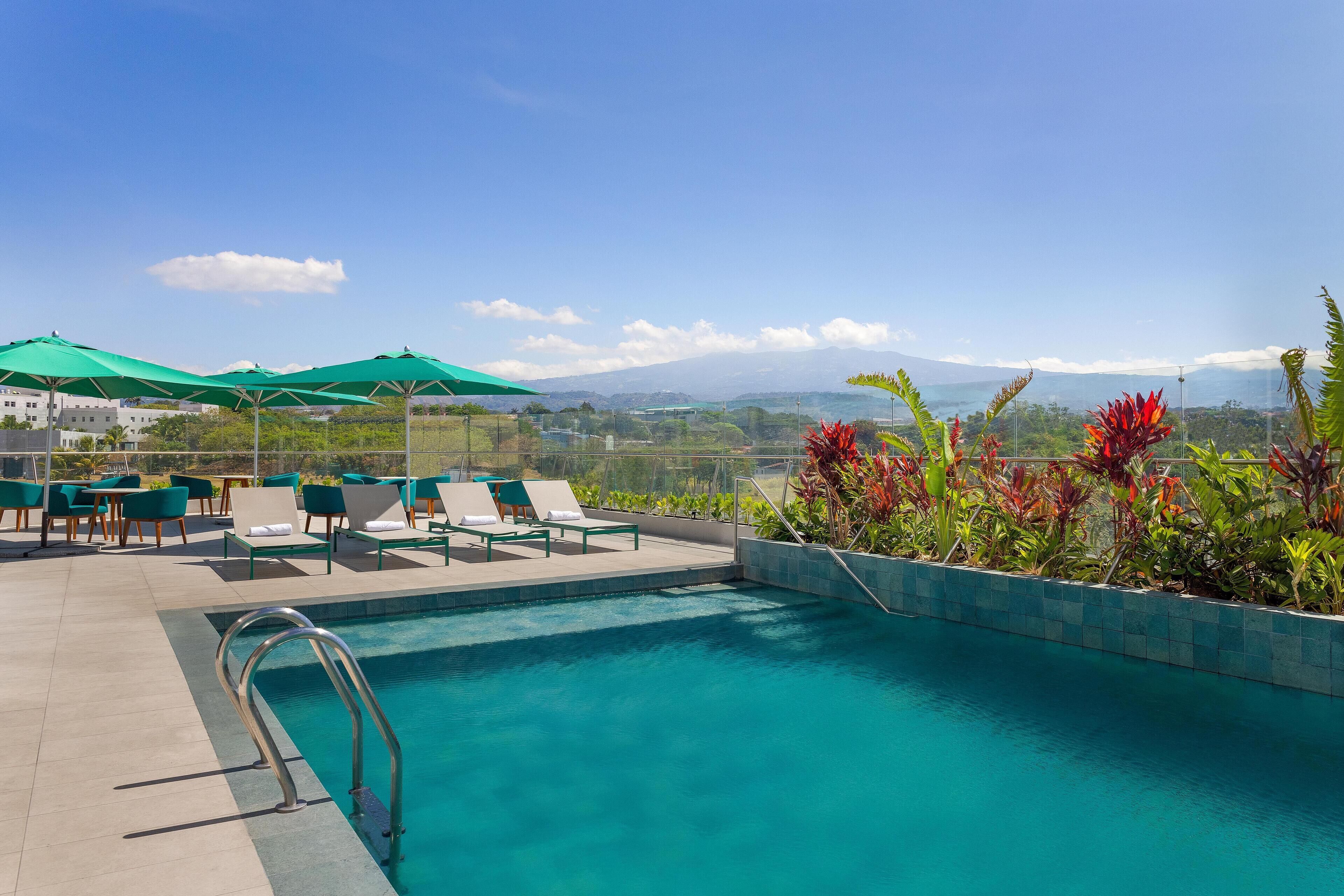 A pool area with lounge chairs and umbrellas, a pool with a view of mountains, and a landscaped area with plants.