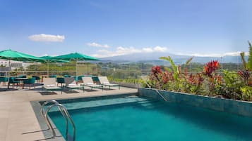 Un área de piscina con sillones de descanso y sombrillas, una piscina con vistas a montañas, y una zona ajardinada con plantas.