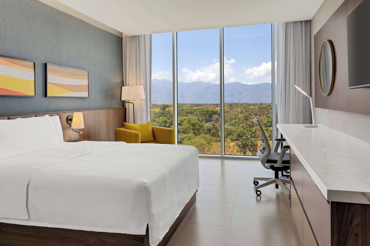A hotel room with a large bed, a desk, a chair, and a view of the mountains.