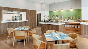 A modern dining area with wooden chairs and tables, a buffet station with various food items, and a green accent wall.