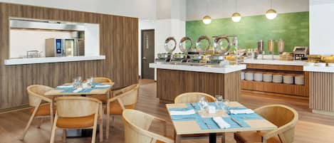 Un área de comedor moderna con sillas y mesas de madera, una estación de buffet con diversos alimentos y una pared de acento verde.