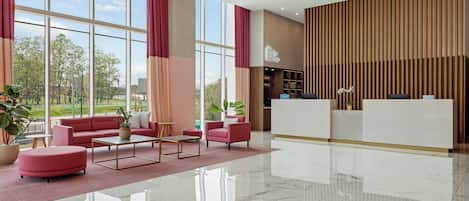 Een moderne hotel lobby met een receptiebalie, een zithoek met banken en poefs, een salontafel, en grote ramen met uitzicht op het buitengebied.
