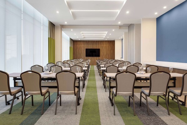 Un salón de conferencias con filas de mesas y sillas, una pantalla grande y piso alfombrado.