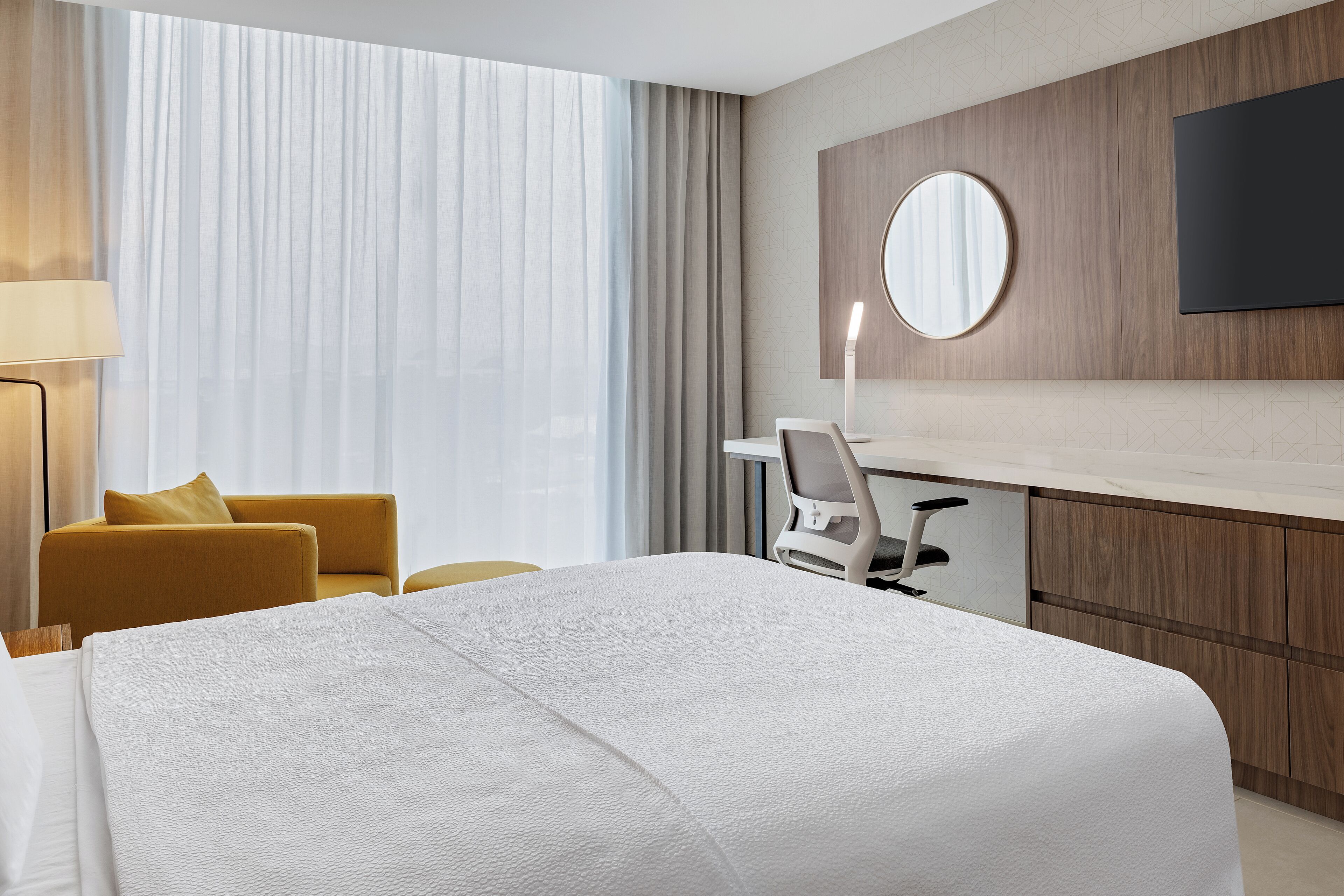 Una habitación de hotel con una cama grande, un escritorio con silla, un televisor montado en la pared y una ventana con cortinas.
