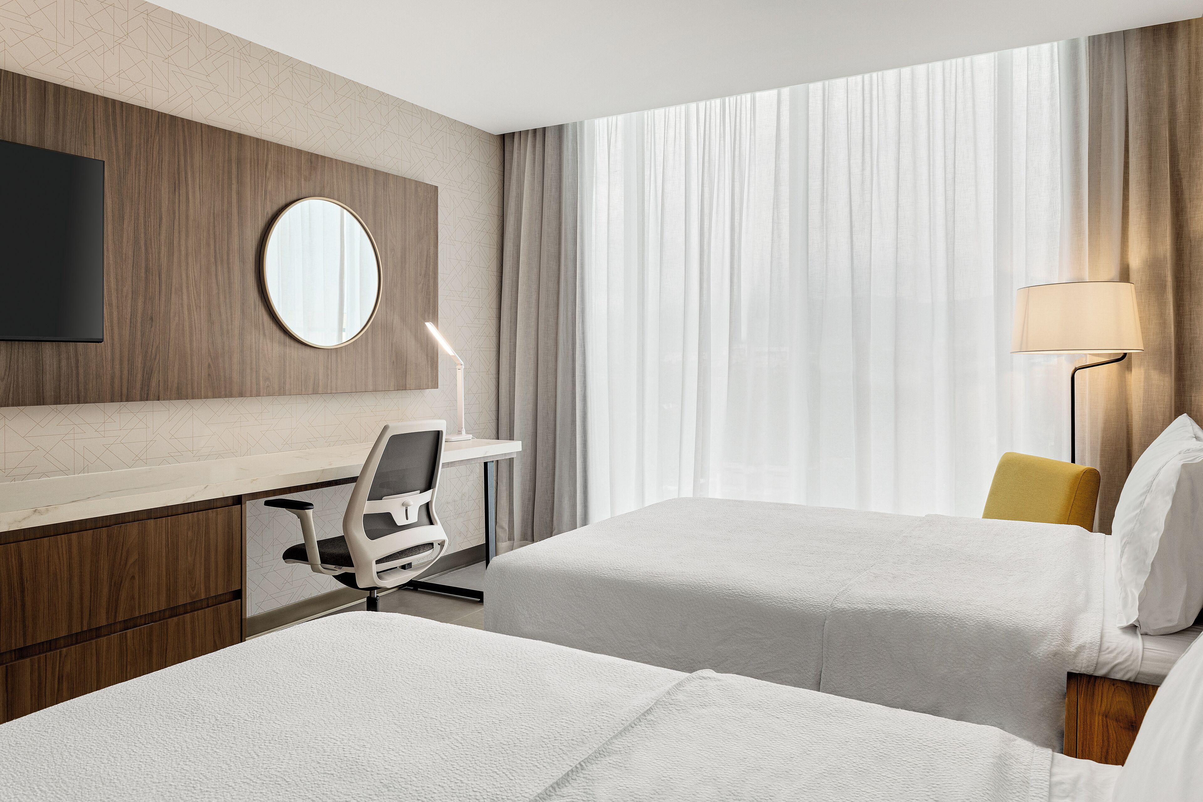 A hotel room with two beds, a desk, a chair, a lamp, and a TV.