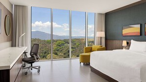 A hotel room with a large window, a bed, a desk, and a chair. - Hilton Garden Inn San Jose Airport City Mall (Alajuela)