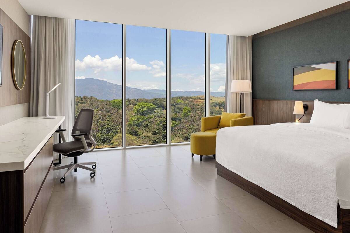 A hotel room with a large window, a bed, a desk, and a chair.