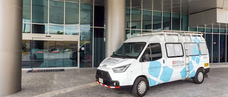 Una camioneta blanca con la marca Hilton Garden Inn estacionada frente a un edificio moderno con grandes ventanales.
