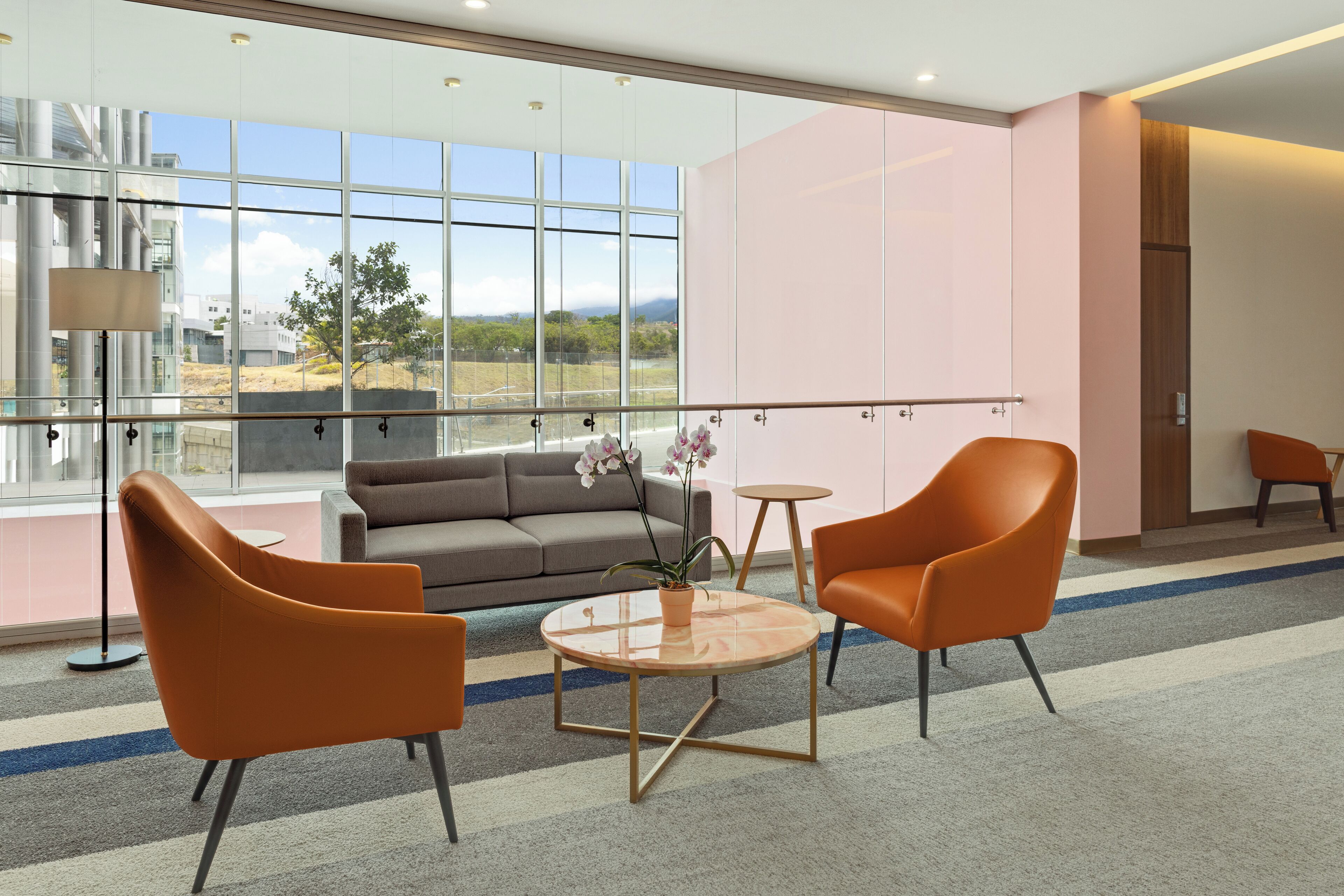 A modern lobby with a sofa, two orange chairs, a small table, and a large window with a view of industrial buildings and a tree.