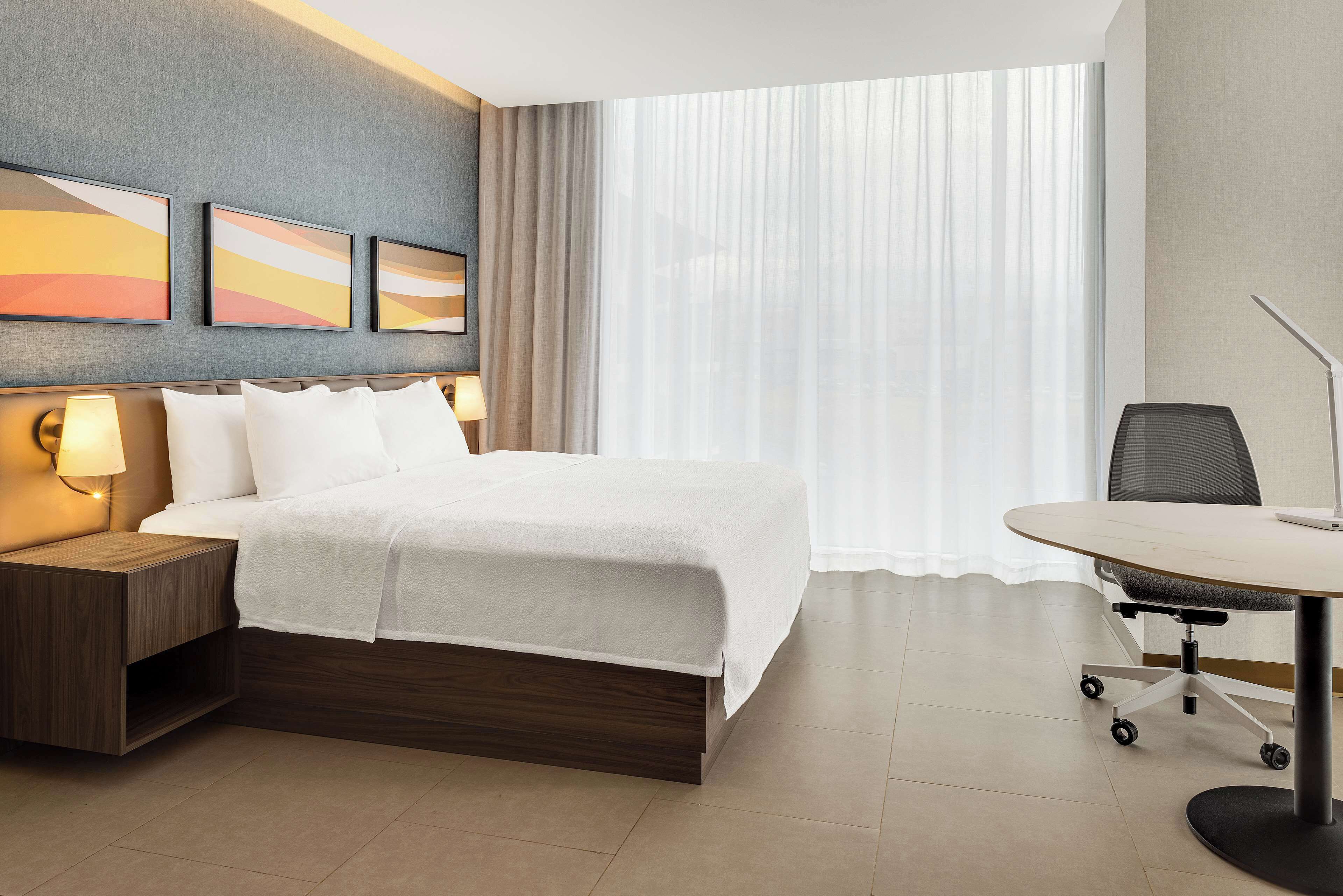 A hotel room with a bed, bedside table with lamp, a desk with chair, and a large window with curtains.