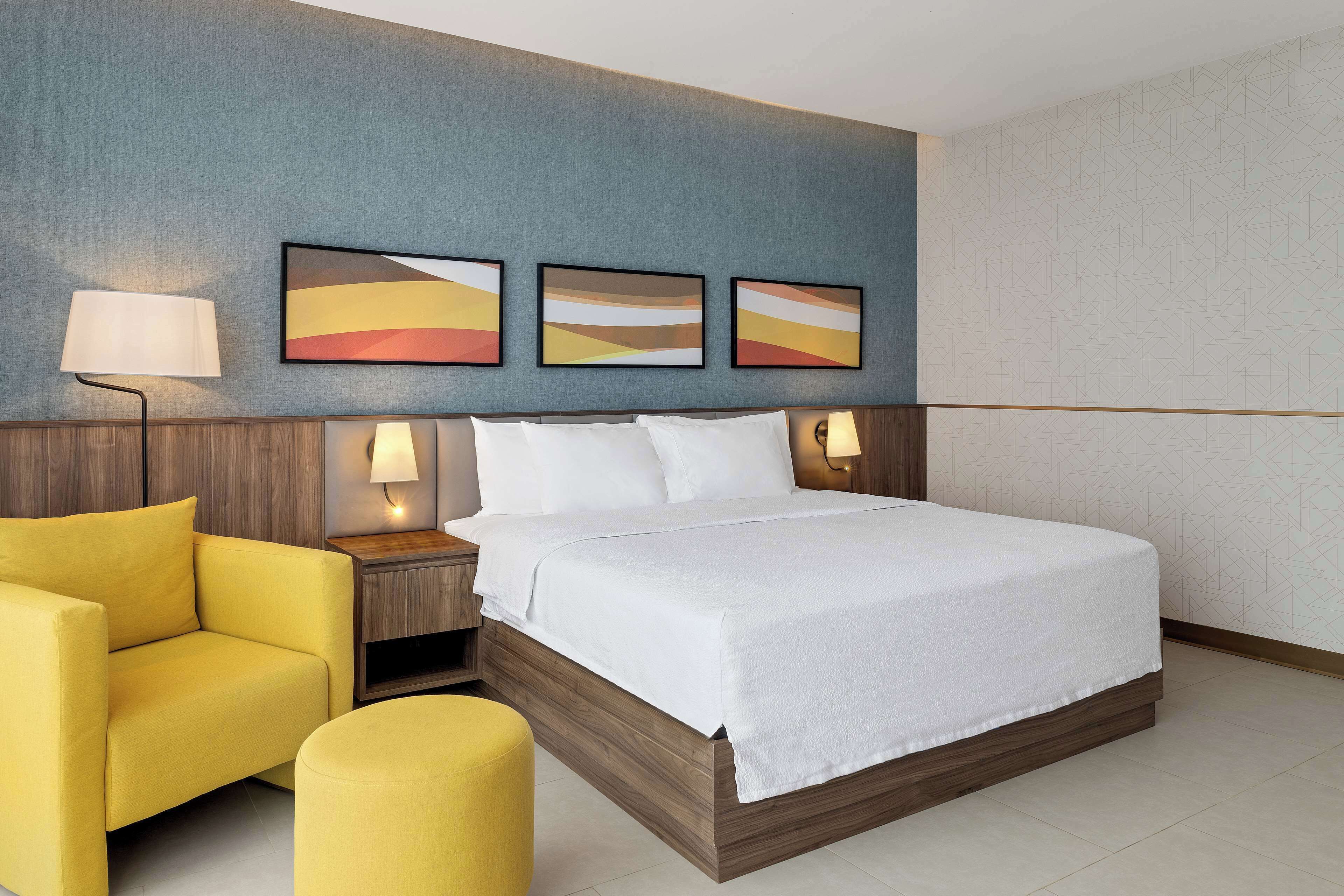 A hotel room with a large bed, a yellow armchair, a bedside table with a lamp, and three abstract paintings on the wall.