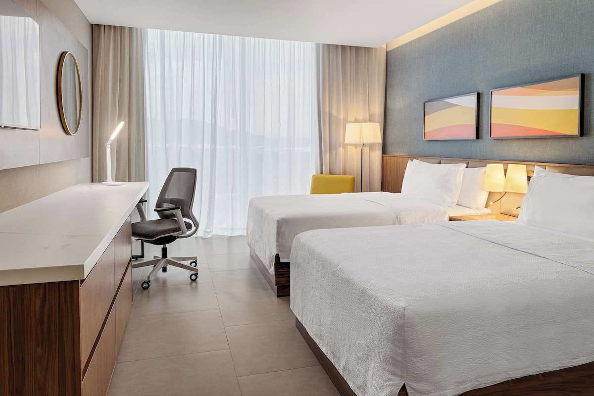A hotel room with two beds, a desk, a chair, and a large window with curtains.
