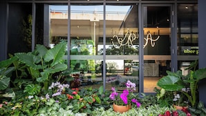 Entrada de hotel con amplios ventanales, jardín con diversas plantas y un interior visible con asientos y un letrero de neón.