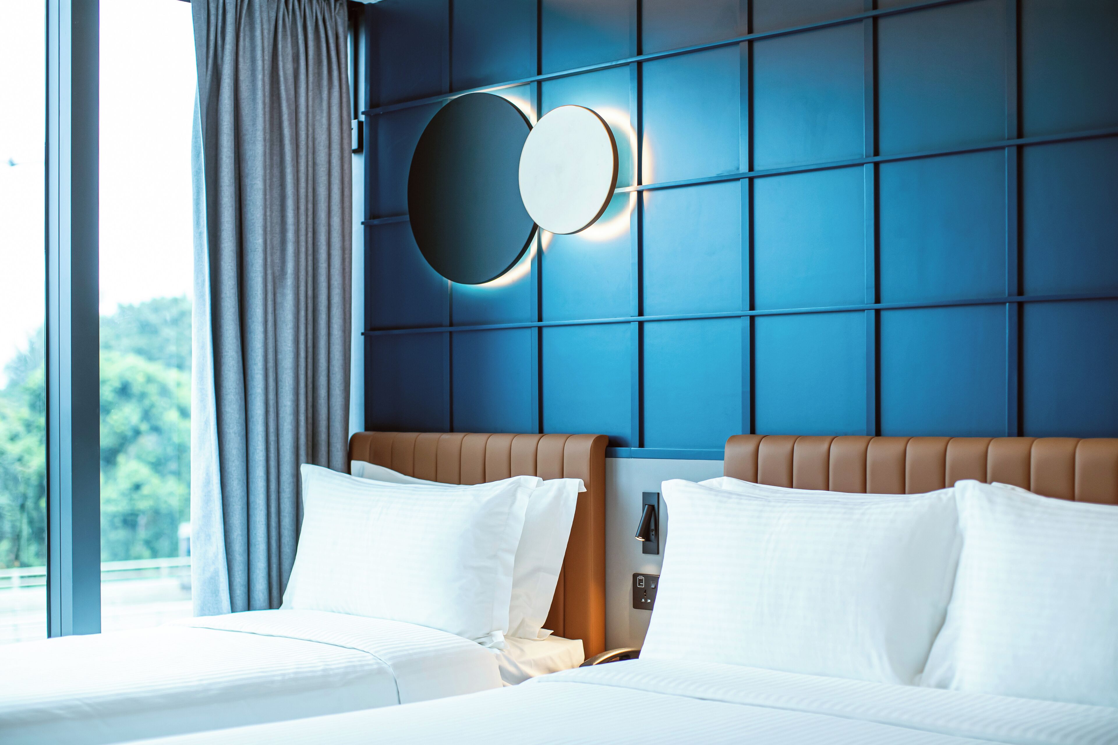 Une chambre d’hôtel avec deux lits, une grande fenêtre avec des rideaux, un mur bleu orné d’un luminaire circulaire, et une vue sur la verdure à l’extérieur.