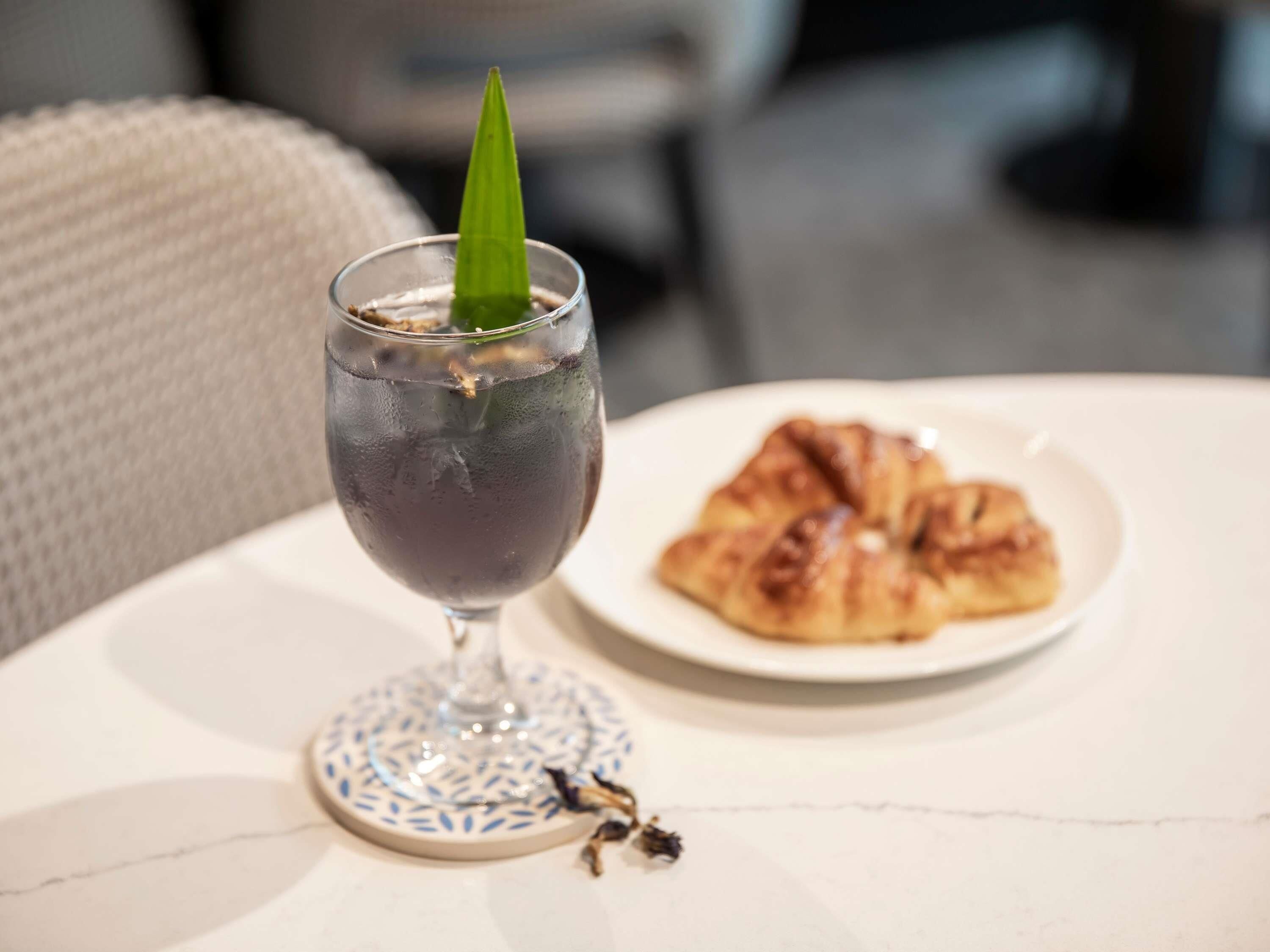 A glass of iced beverage with a green leaf garnish on a coaster, a plate of croissant, and a small bunch of dried flowers on a table.