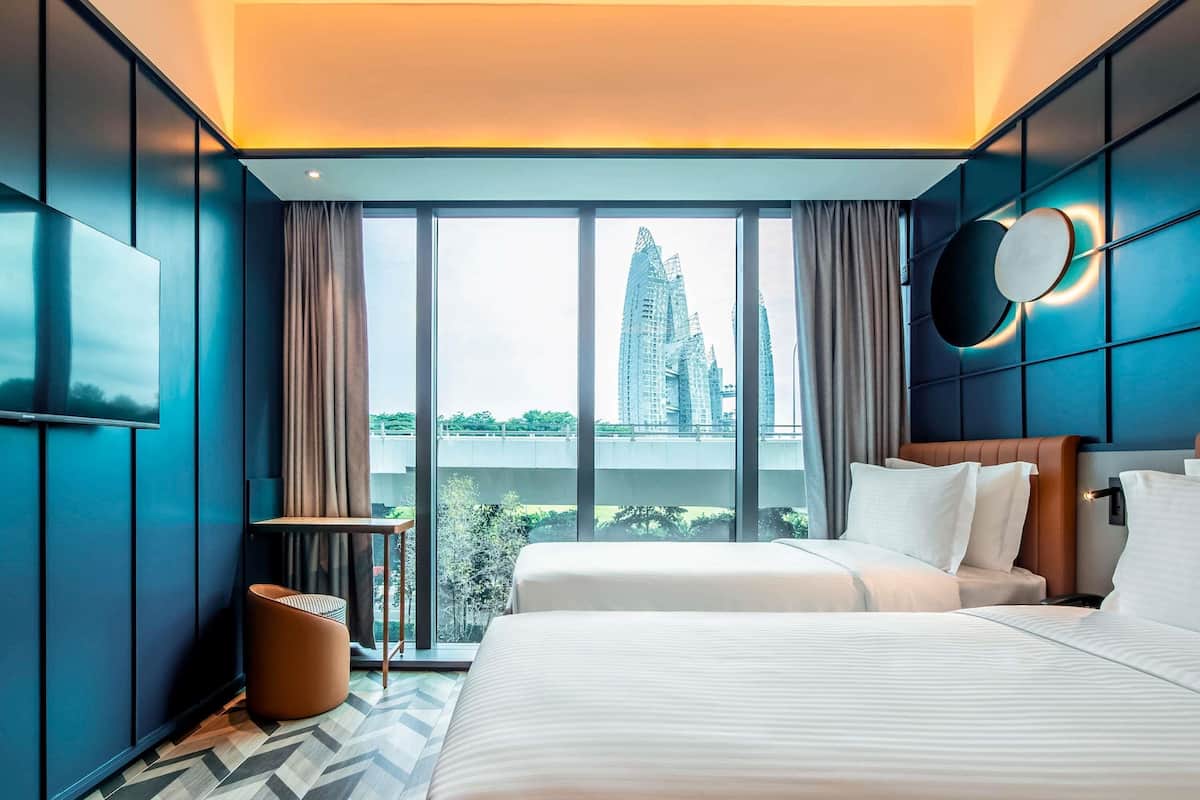 Habitación de hotel moderna con una cama grande, un pequeño espacio para sentarse y vistas a un paisaje urbano a través de amplios ventanales.