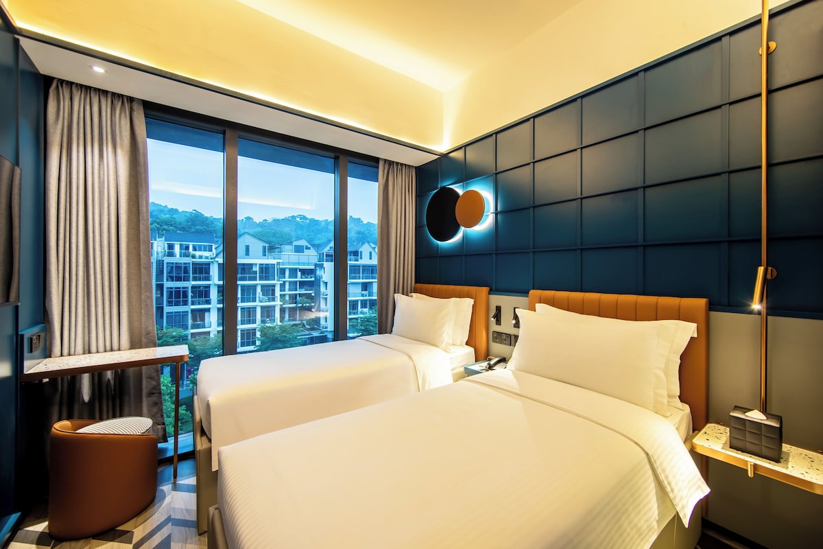 A hotel room with two beds, a large window with a view of buildings, a round wall light, and a nightstand with a lamp.