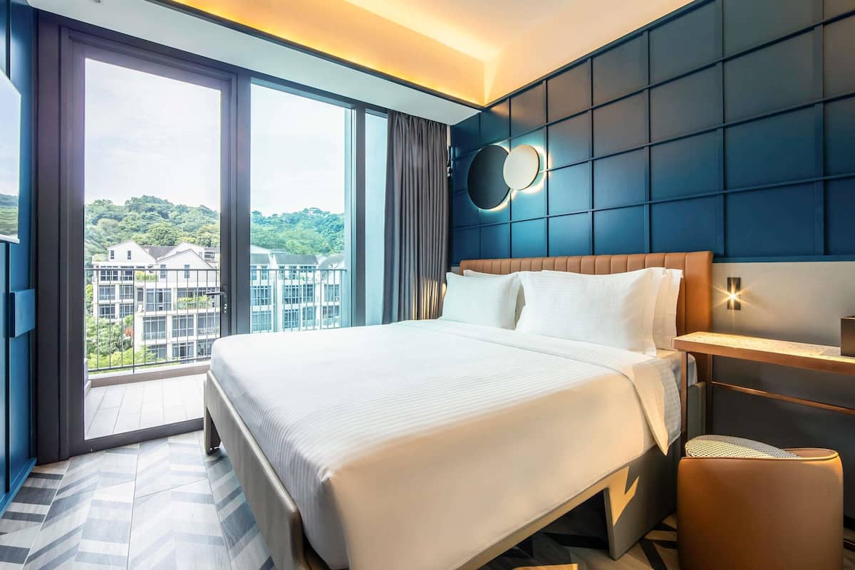 Habitación de hotel moderna con una cama grande, una mesita de noche, un espejo redondo y vistas a edificios y zonas verdes.