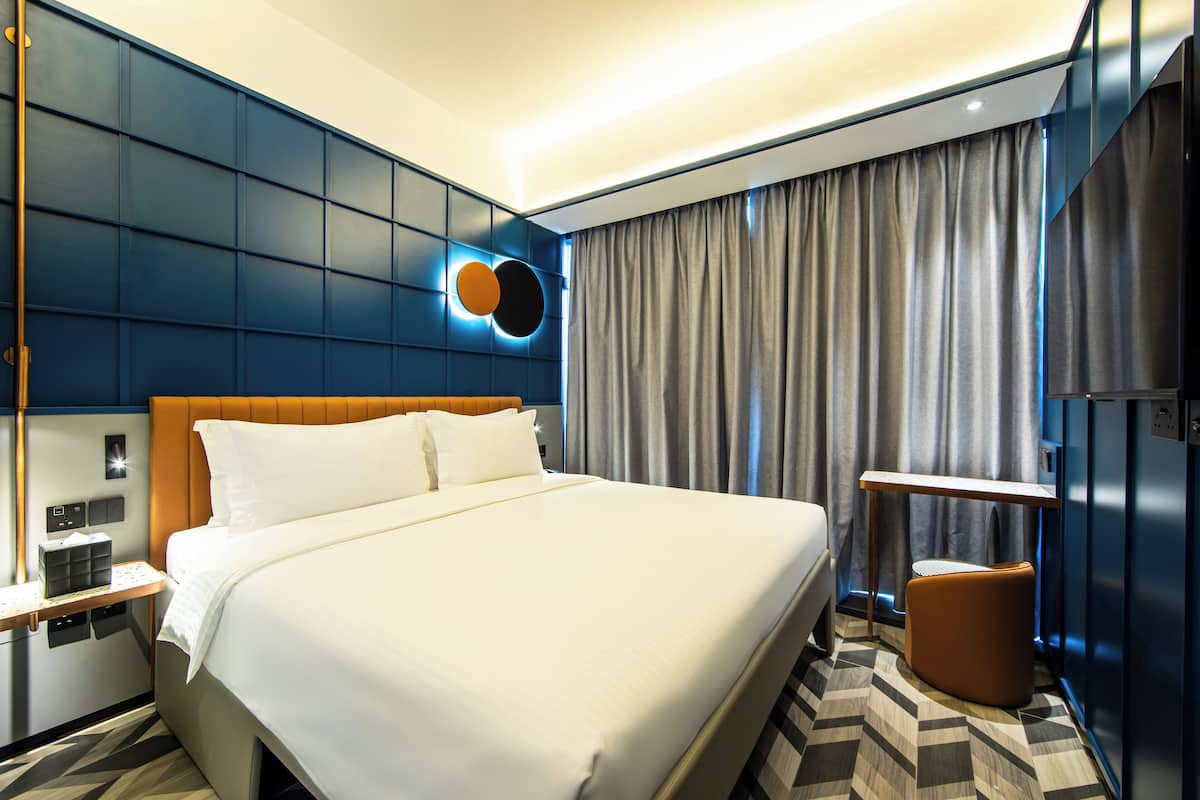 Una habitación de hotel moderna con una cama grande, una mesita de noche, un pequeño rincón de descanso y una lámpara de pared.