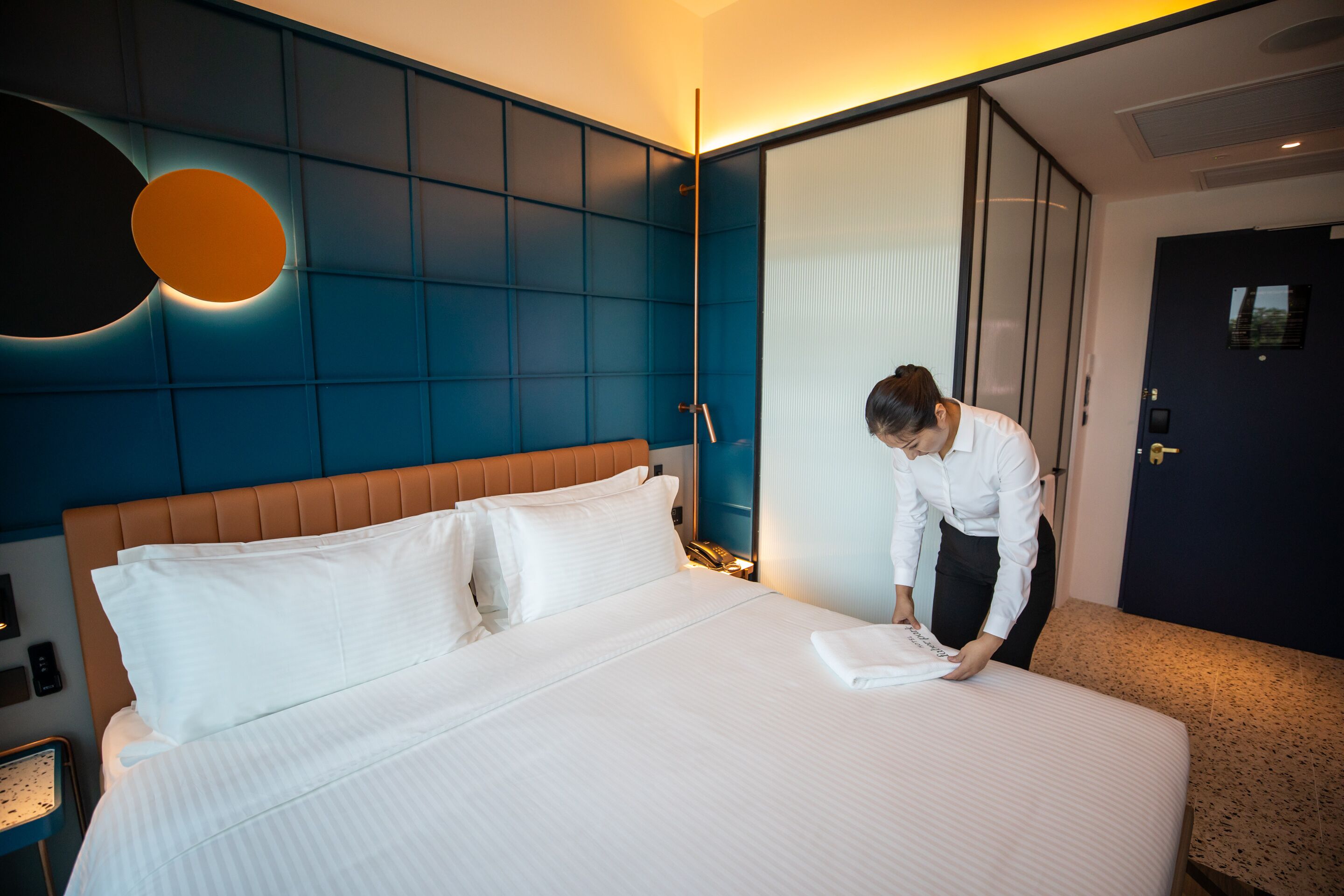 A hotel room with a large bed, a wall-mounted circular light, a wardrobe, and a door with a lock.