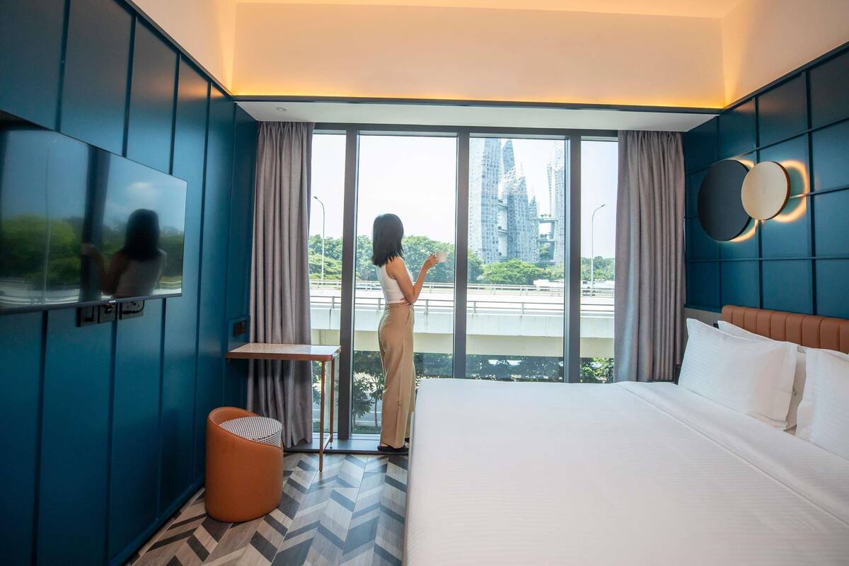 A hotel room with a large bed, a TV mounted on the wall, a small table, and a view of a cityscape through large windows.