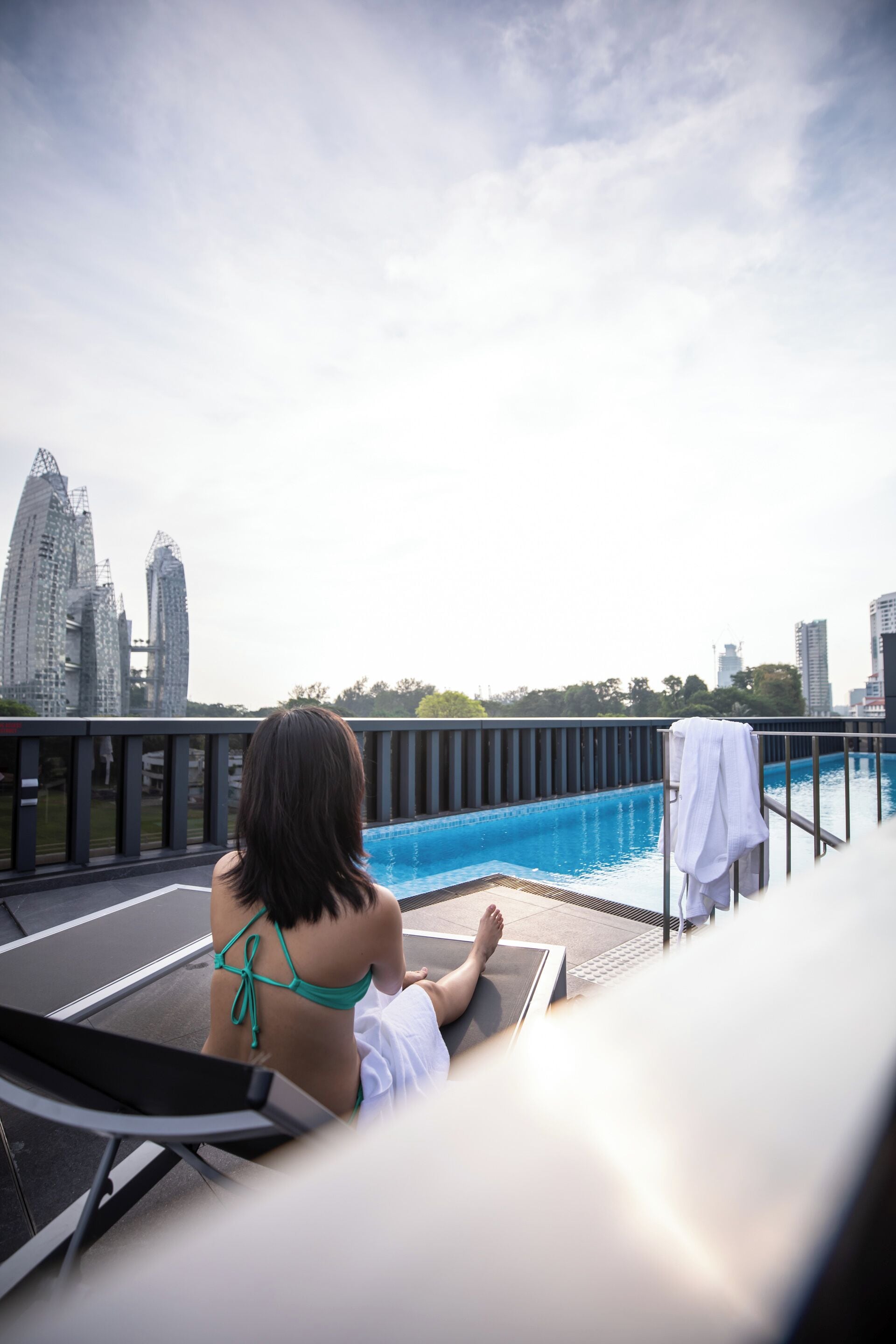 A woman in a green bikini sitting by a pool with a cityscape in the background.