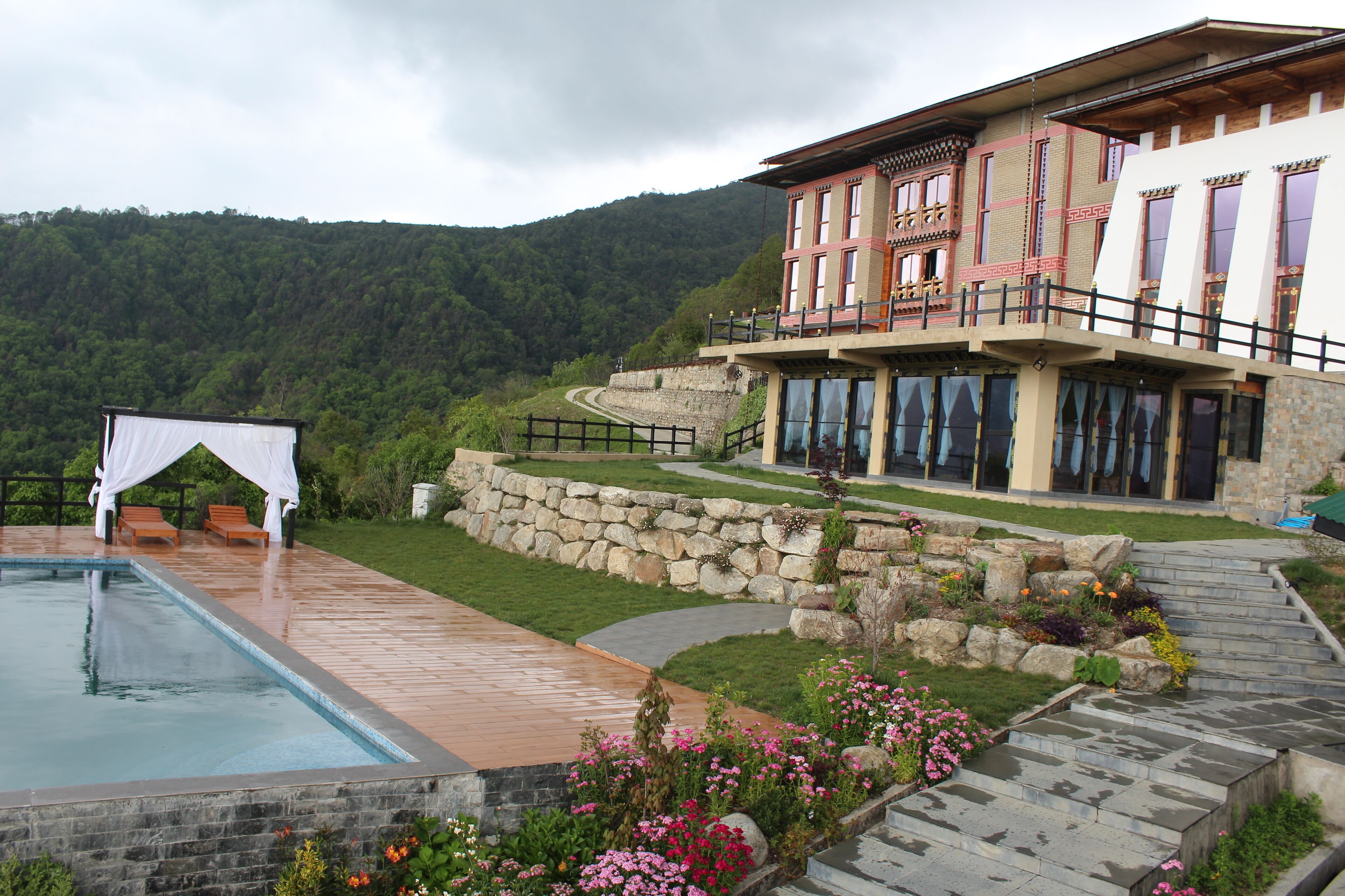 A pool with a wooden deck, a stone wall, and a flower bed with pink flowers.