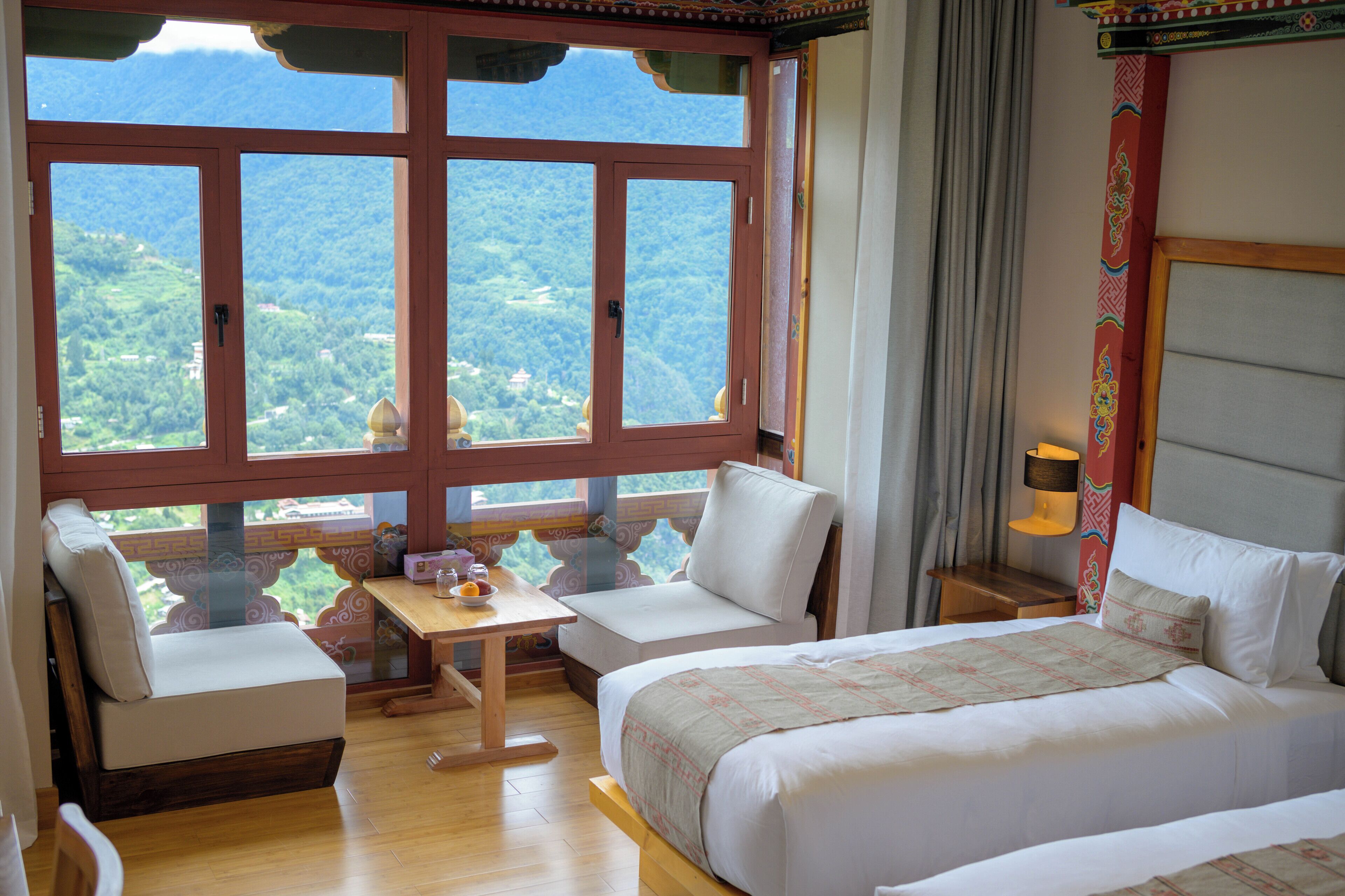 Ein Hotelzimmer mit großem Fenster und Bergblick, einem Bett mit gemustertem Überwurf, einem kleinen Tisch mit Obstschale und einem weißen Sofa.