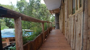 Una terraza de madera con barandillas, rodeada de follaje y una edificación.