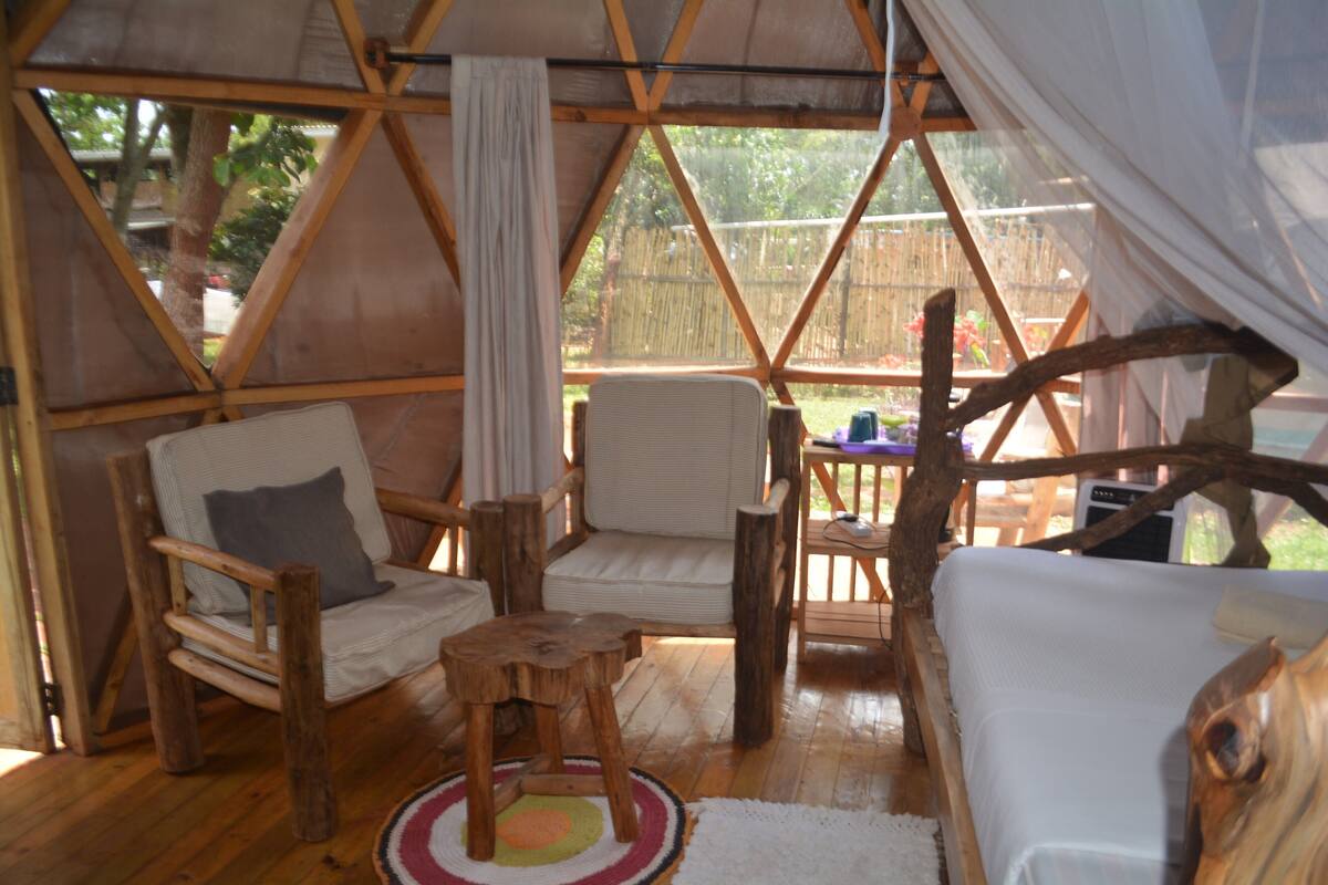A cozy interior with a wooden floor, two cushioned chairs, a small wooden stool, and a bed with a white cover.