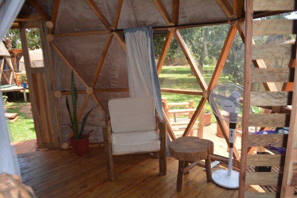 A wooden tent with a cushioned chair and a small table, a fan, and a potted plant.